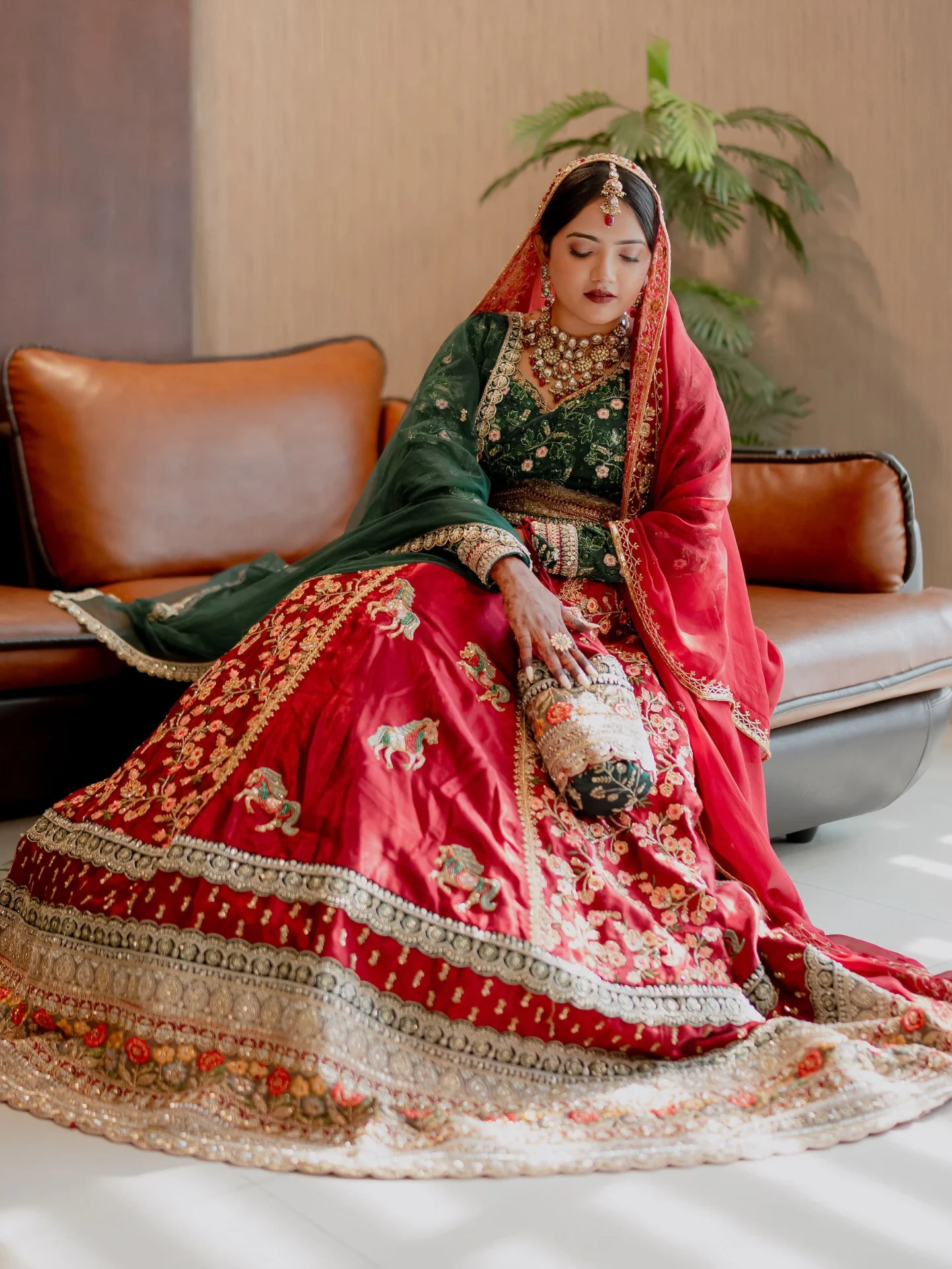 A bride in a deep red and gold lehenga seated against a leather couch